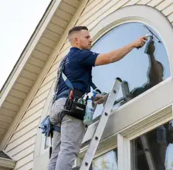 Professional window cleaner on a ladder using a squeegee to clean an exterior arched window, Tempe, AZ.